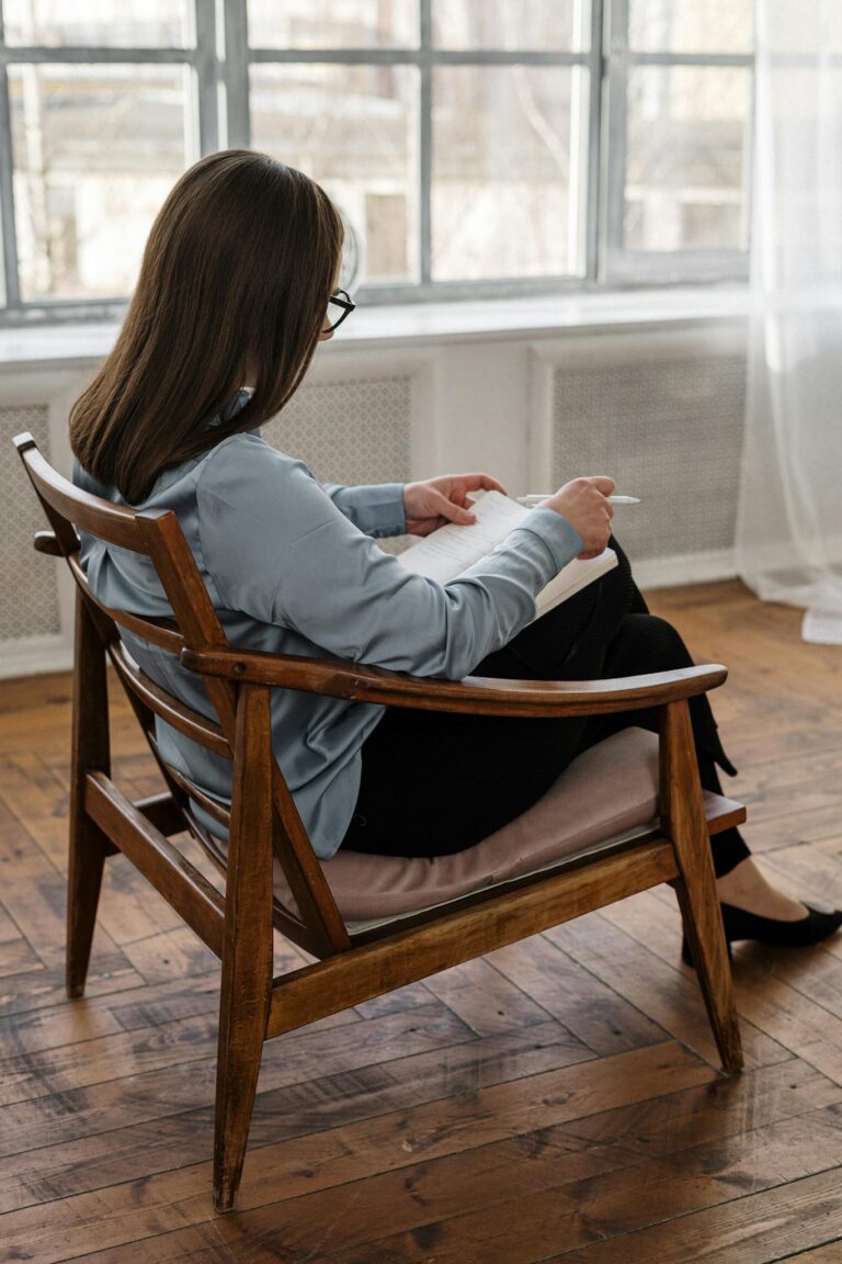 Woman with glasses sitting in a wooden chair while writing in a notebook, capturing thoughtful moments.